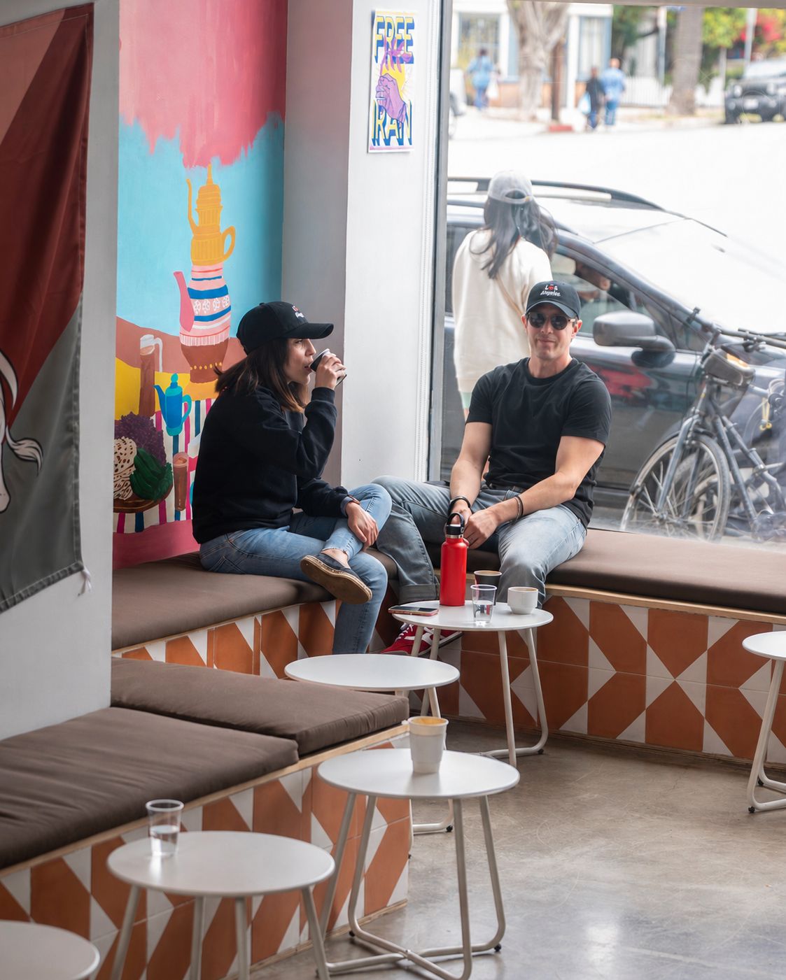 A couple on the tiled window bench at Caspian Coast Coffee, Venice, with the Persian teapot mural and Free Iran poster